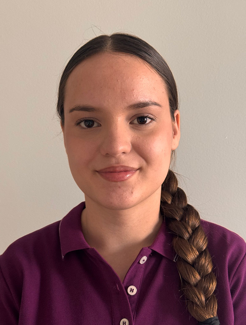 Eine junge Frau mit freundlichem Gesichtsausdruck, braunen Haaren, lila Hemd vor hellem Hintergrund.